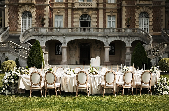 Reception tablescape with an outdoor wedding reception table, ivory linens, taper candles, white florals and greenery before a brick mansion terrace