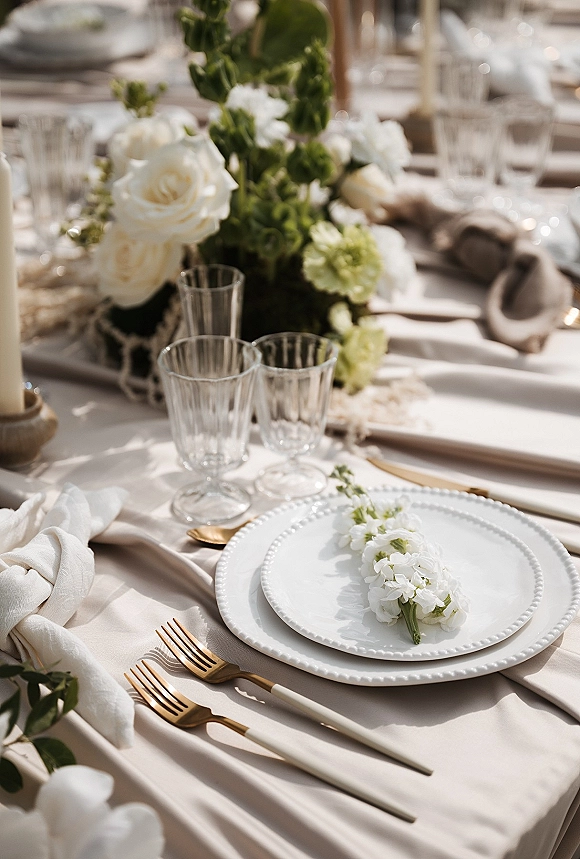 Reception tablescape with a wedding place setting, white and green centerpiece, gold flatware, clear goblets, and ivory taper candles outdoors