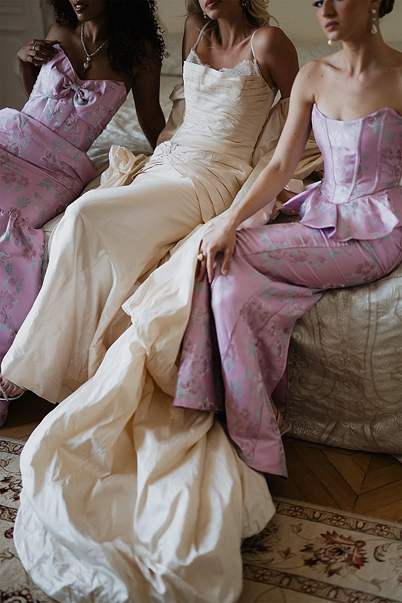 Bridal party portrait of bride with bridesmaids sitting on a bed, wearing corset gowns and pink floral dresses with lace trim accents