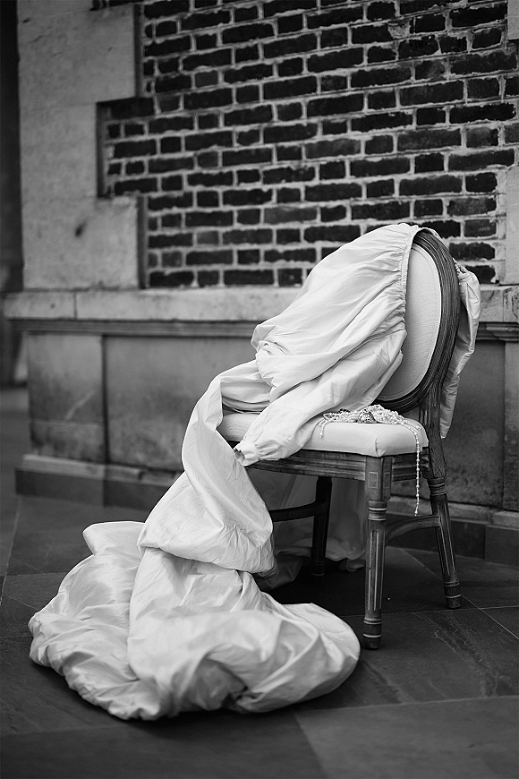 Wedding dress detail with gown draped on antique chair, long train on stone floor and pearl necklace accents against brick wall indoors
