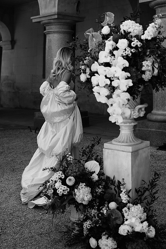 Bridal portrait in a black and white wedding portrait style, bride walking away in a gown with long train beside a large urn of orchids in a stone-column courtyard