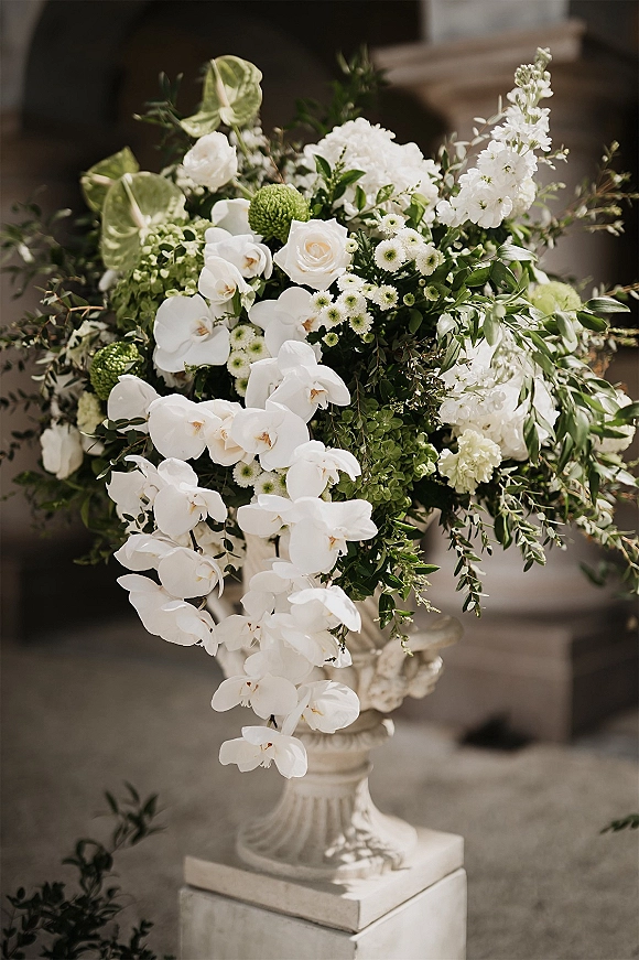 Wedding floral arrangement, ceremony urn flowers with cascading white orchids and roses on a stone urn pedestal by stone steps outdoors