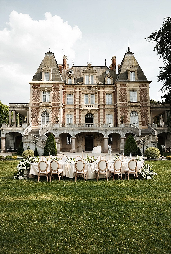 Outdoor reception setup with a long banquet table, white tablecloth, floral centerpieces and taper candles on a stone terrace before a mansion staircase