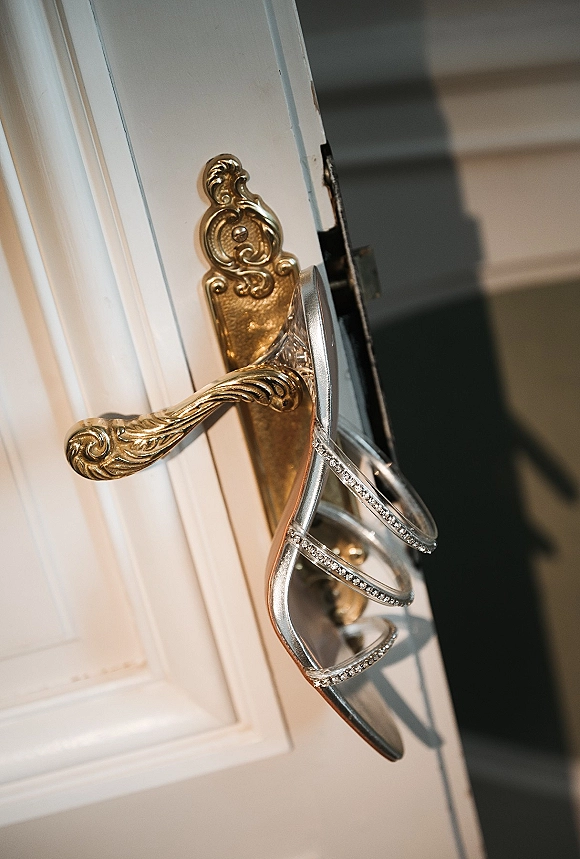 Wedding shoes, silver wedding heels with rhinestone straps hanging from an ornate brass door handle on a white hallway door by stairs