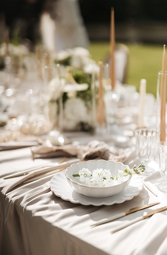 Reception tablescape with scalloped plates, white floral centerpiece and tapered candles on a lawn, with soft greenery and blurred guests behind