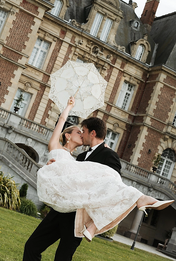 Wedding kiss portrait of groom carrying the bride in a tuxedo and strapless dress, holding a lace parasol on a mansion stone staircase
