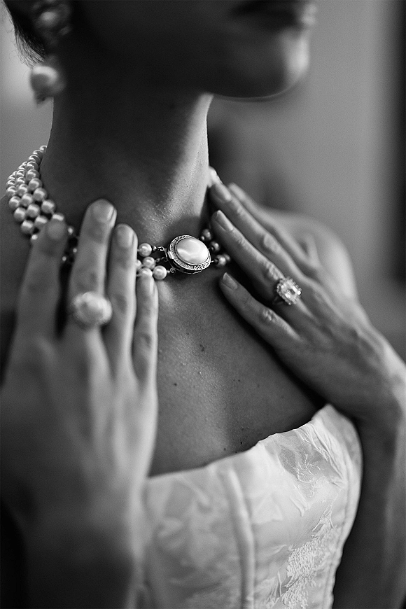 Bridal jewelry close-up of a pearl necklace bride adjusting a pendant at her collarbone, engagement ring visible above a strapless lace bodice indoors