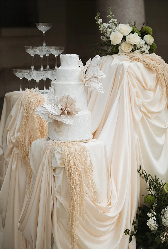Wedding cake with textured buttercream on a draped table, sugar flowers and white roses beside a champagne coupe tower against a stone wall