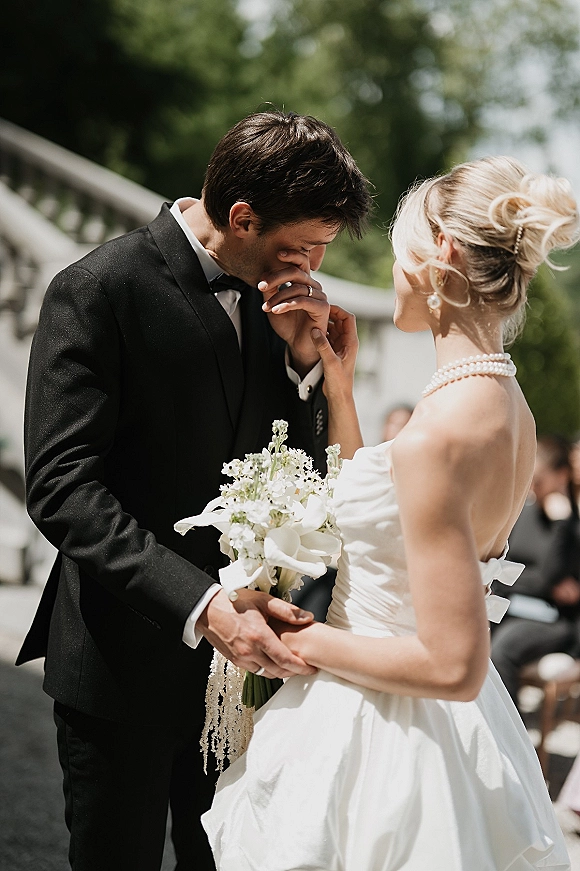 Ceremony moment as groom kisses bride’s hand while they hold hands, her strapless gown and bouquet on stone steps before guests