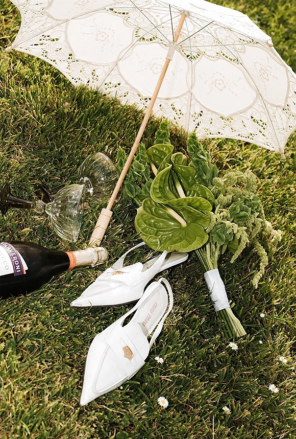 Wedding details flat lay with lace parasol, bouquet, champagne bottle, wine glasses, and white slingback heels arranged on grass lawn