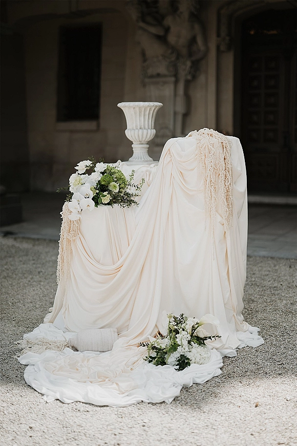 Ceremony pedestal decor with draped fabric and white florals with greenery in an urn by a stone doorway in a gravel courtyard