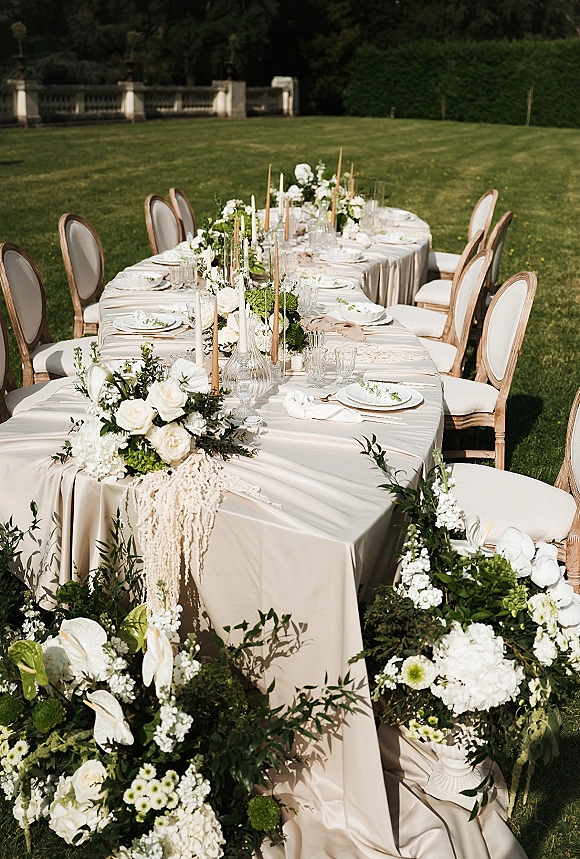 Reception tablescape with an outdoor wedding dinner table on a lawn, draped ivory linen, white roses and hydrangea garland, taper candles in glass.
