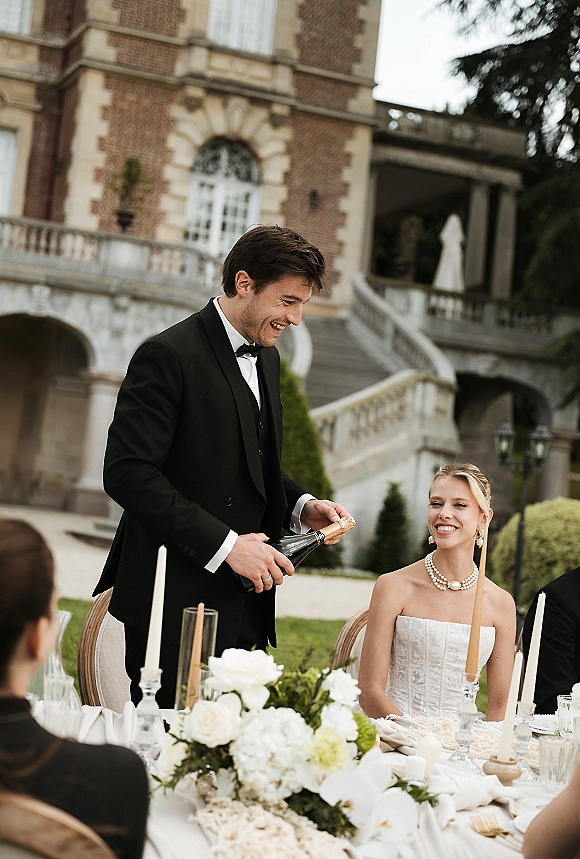 Wedding toast as groom pours champagne beside bride in strapless dress and pearls at candlelit terrace table before a historic mansion