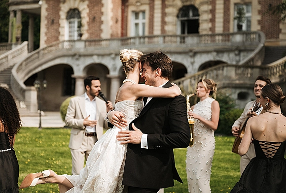First dance outdoors as groom lifts bride in strapless dress with pearl necklace, live wedding band on estate lawn by historic facade