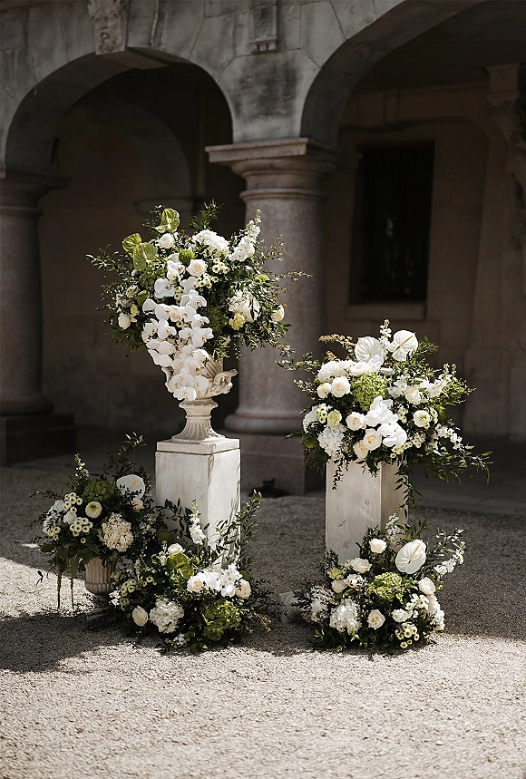 Ceremony floral arrangements of white blooms and greenery in urns on plinths with ground clusters against stone arches in a courtyard