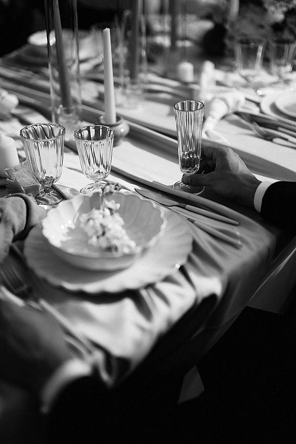 Reception tablescape with wedding place setting, taper candle centerpiece, glass stemware, folded napkins, and floral runner as guests blur behind