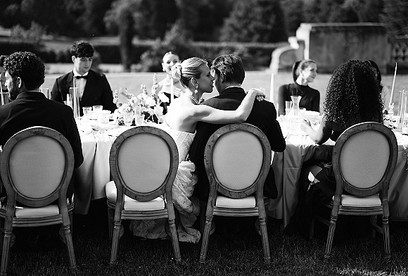 Reception moment as the bride hugging groom at the head table, surrounded by long taper candles and floral centerpiece on a garden lawn