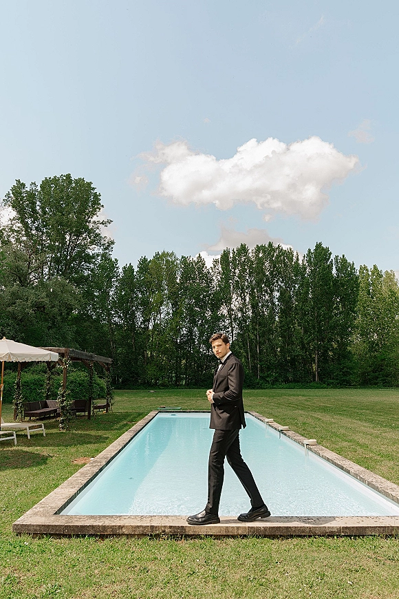 Groom portrait in a classic black tuxedo with bow tie and dress shoes, posing poolside on a lawn under blue sky near a pergola
