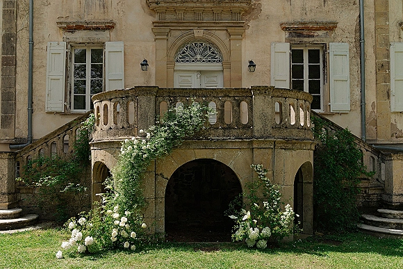 Wedding ceremony backdrop with white floral arrangements and climbing greenery on a stone mansion facade with balcony and staircase
