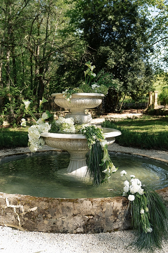 Wedding fountain decor with a fountain floral arrangement of white calla lilies, hydrangeas, and greenery garland on a stone tiered fountain in a garden setting