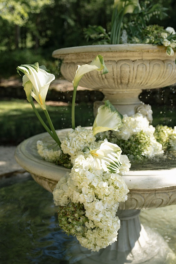 Fountain floral decor with wedding fountain flowers, white calla lilies and hydrangeas draped in greenery garland on a sunlit stone tiered fountain