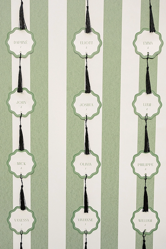 Wedding seating chart with hanging escort card display featuring scalloped green name tags with black tassels against a green-and-white striped backdrop