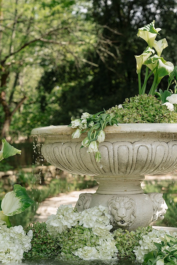 Wedding fountain decor with white calla lilies and hydrangeas spilling over a stone fountain, framed by lush garden trees and greenery