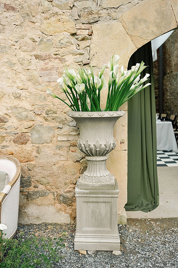 Wedding floral urn with white calla lilies on a stone pedestal, set by an arched doorway with green drapery and stone wall backdrop