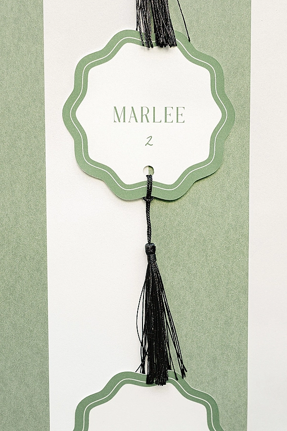 Wedding place card with die-cut tag, green border, and black tassel on string, styled on layered white and green paper flat lay