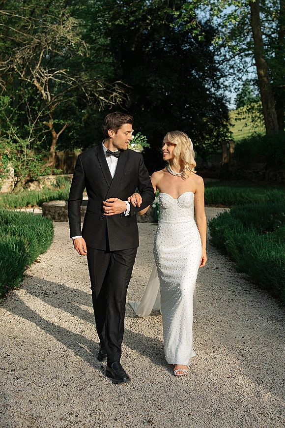Couple portrait of bride and groom walking arm in arm on a sunlit garden path, her strapless beaded dress and his tuxedo visible