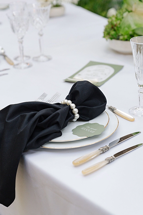 Reception tablescape with a black napkin and pearl napkin ring on gold-rimmed plates, crystal flutes, and a floral centerpiece amid greenery