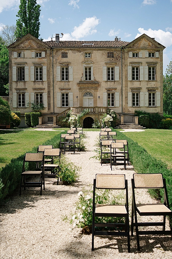 Outdoor ceremony setup with garden wedding ceremony seating, wood and cane chairs flanking a gravel aisle lined with white florals and greenery, villa facade behind