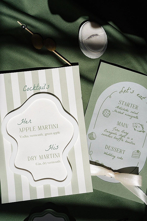 Wedding menu cards with wedding cocktail menu card tied in silk ribbon, scalloped labels and stirrers on green fabric with shadows