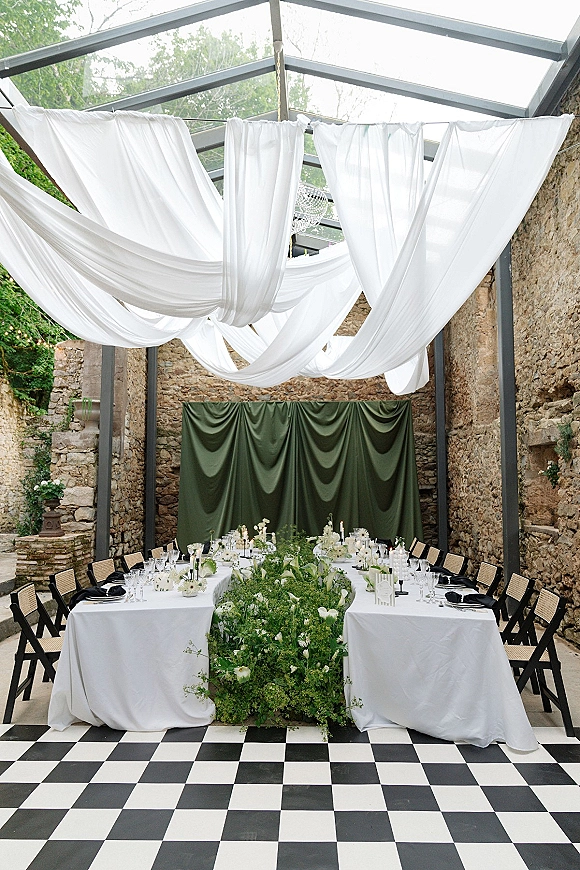 Reception tablescape with U-shaped head table, greenery runner, white florals and black taper candles under a draped ceiling and chandelier