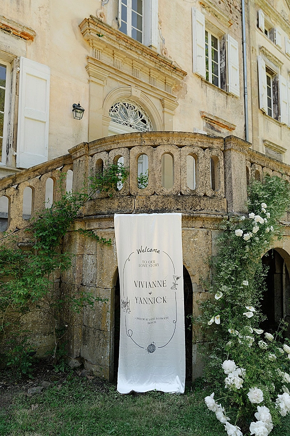 Wedding welcome sign with calligraphy on a hanging fabric banner, framed by white flowers and greenery on a stone terrace entry