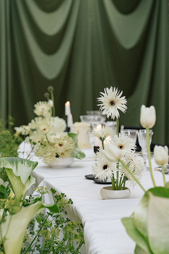 Reception tablescape with white wedding tablescape details, bud vases of tulips and calla lilies, taper candles, and dark green drapery backdrop