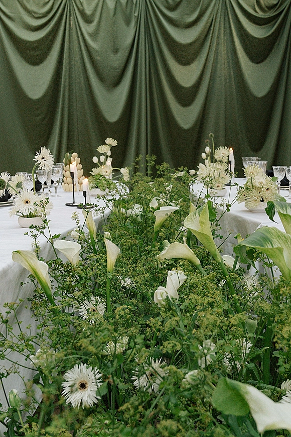 Reception tablescape with a green and white tablescape, calla lilies and greenery garland, taper candles, and glassware before a green draped curtain backdrop