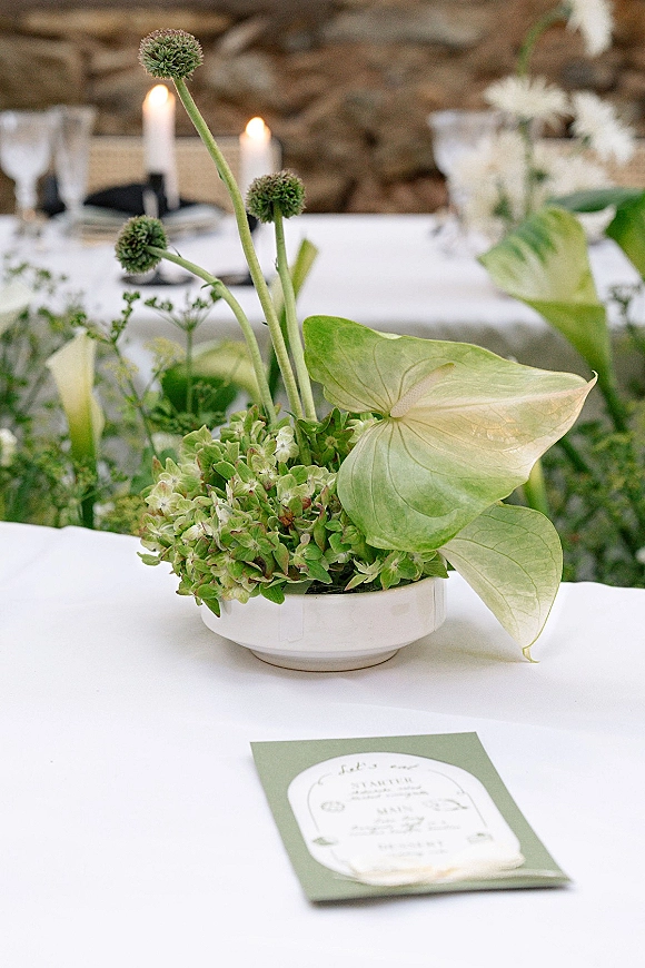 Wedding centerpiece with green wedding centerpiece florals—hydrangea, anthurium leaves, and allium stems with taper candles on a white tablecloth by a stone wall