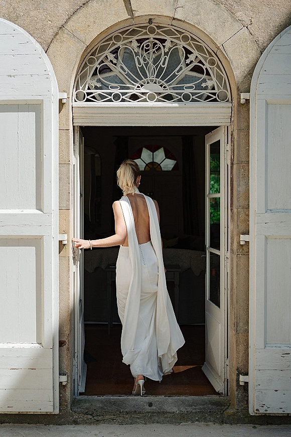 Bridal portrait of a bride in a backless wedding dress with draped cape train stepping into an arched doorway by white shutters and stained glass