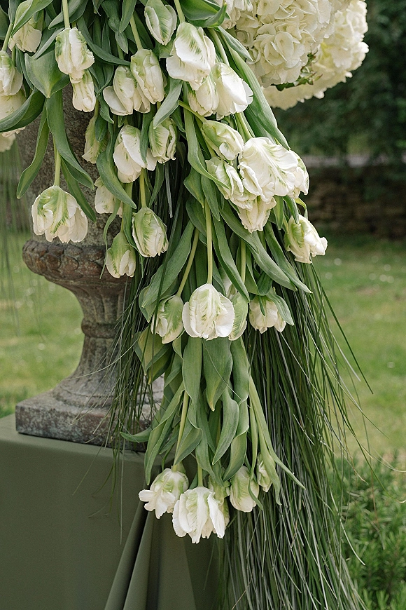 Wedding floral arrangement of white tulip wedding flowers cascading with greenery from a stone urn on linen, set on a garden lawn with trees