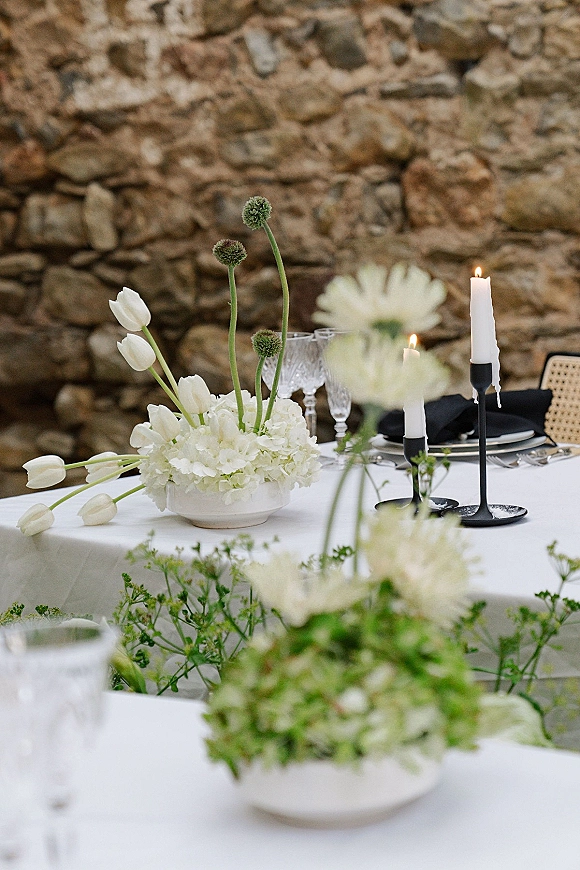 Reception tablescape with a white wedding tablescape of tulips and hydrangea centerpiece, taper candles, and black napkins by a stone wall