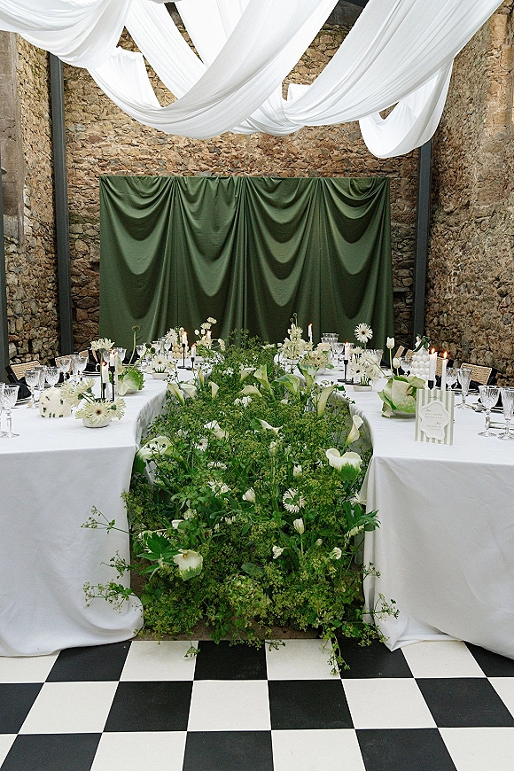 Reception tablescape with wedding head table decor featuring white linens, floral runner, taper candles, and glassware on a checkered floor