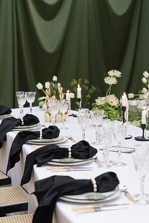 Reception tablescape with a black and white wedding table, pearl napkin rings, crystal stemware, taper candles, and green drapery backdrop