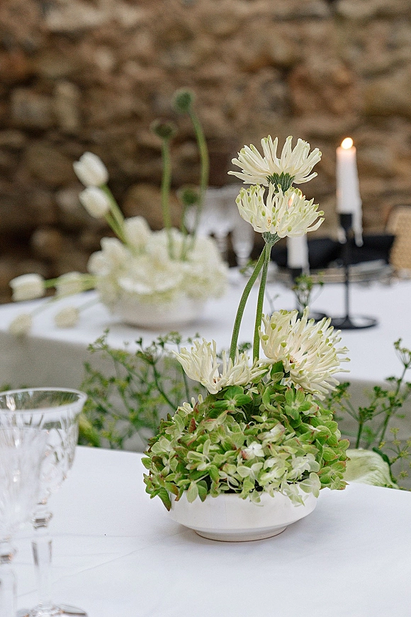 Wedding table centerpiece with low floral centerpiece of white flowers and greenery in a bowl vase, flanked by black taper candles on a stone-wall table