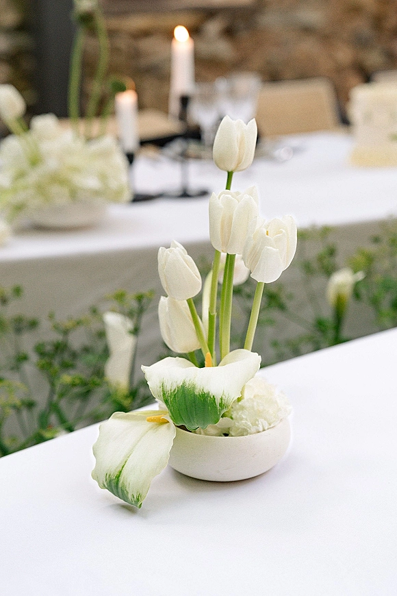 Wedding centerpiece with tulip wedding centerpiece styling, white tulips and calla lilies in a low bowl beside taper candles on a white tablecloth
