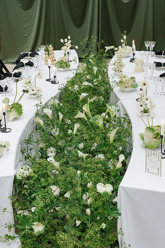Reception tablescape with a u shaped wedding table, white linens, calla lilies, candles, crystal glassware, and a green draped backdrop