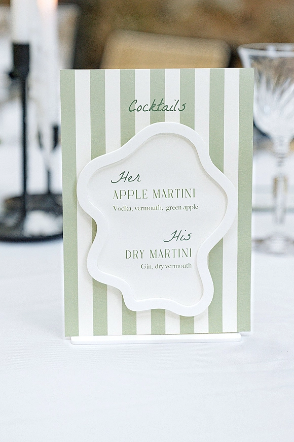 Wedding cocktail sign with green and white striped cocktail menu card in a stand beside glassware on a white linen table