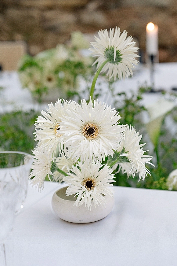Wedding centerpiece with white floral centerpiece in a bud vase, greenery runner, and a white taper candle on a linen table by a stone wall