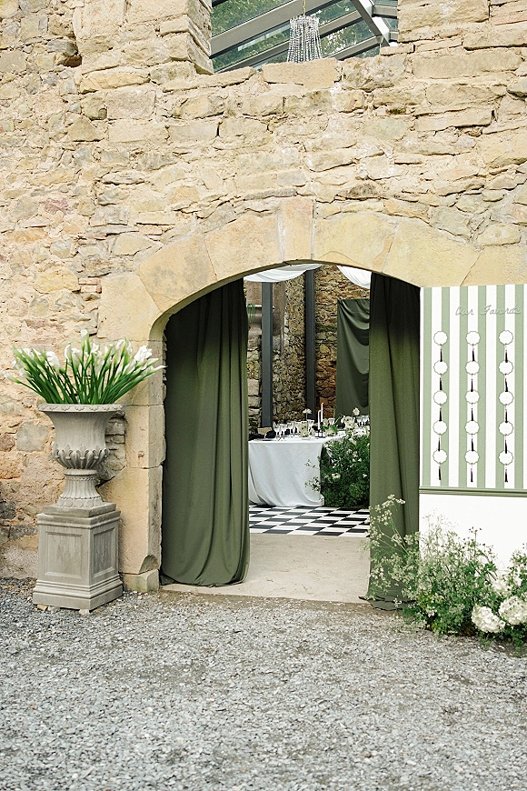 Reception entrance decor with draped entryway curtains, green swag and white floral urns under a stone archway, candles leading to tables inside