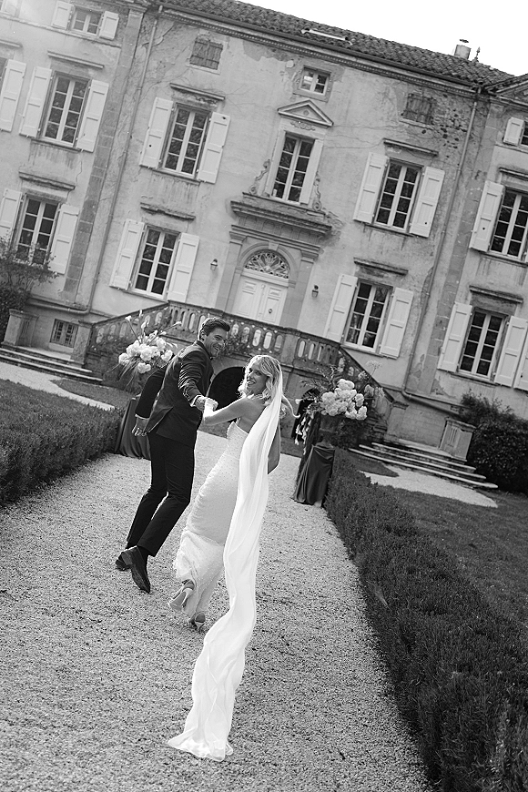 Wedding couple portrait of bride and groom holding hands walking away, long veil train flowing on a gravel path by a historic estate facade